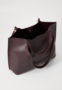 Sac fourre-tout en cuir bordeaux avec deux longues poignées, ouvert en haut, texture lisse, quincaillerie dorée et poche intérieure.