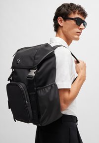 Black nylon backpack with a top flap, zippered front pocket, and logo detail. Features padded shoulder straps and sleek, minimalistic design.