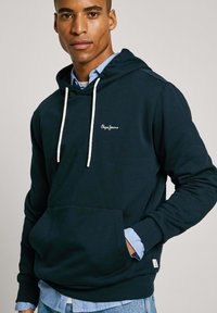 Sudadera oscura azul marino con un bolsillo frontal, cordones blancos y un logo bordado. Superpuesta sobre una camiseta de color azul claro, textura de mezcla de algodón.