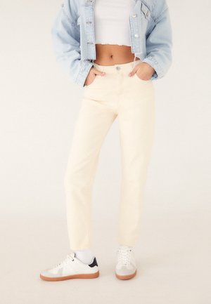 Persoon draagt een lichtblauwe cropped spijkerjas, een witte cropped tanktop, crèmekleurige hoge taille broek, witte sokken en witte sneakers met gumzolen.