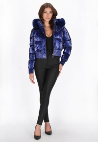 Krótkia niebieska kurtka puchowa z błyszczącym wykończeniem i kapturem z sztucznego futra. Ściągacze na mankietach i dole, z przodu zamek błyskawiczny, zestawiona z czarnymi legginsami i szpilkami.