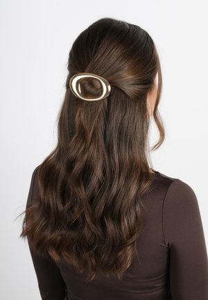 Barrette ovale en or maintenant des cheveux bruns ondulés, avec une texture lisse et une finition polie. Design minimaliste ; positionnée près de la couronne.