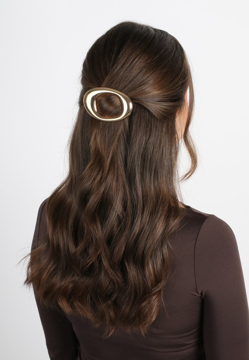 Clip per capelli ovale dorato che tiene in posizione capelli castani mossi, con una texture liscia e una finitura lucida. Design minimalista; posizionato vicino alla corona.