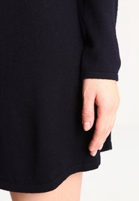 Abito lavorato a maglia blu navy con maniche lunghe, caratterizzato da una texture liscia e un orlo sottile, con una mano posata sul tessuto.