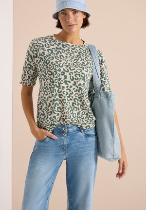 Jonge vrouw draagt een groen en beige luipaardprint top, lichtblauwe spijkerbroek, blauwe bucket hat en draagt een lichtblauwe tas.