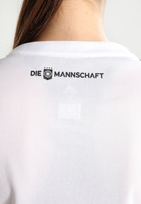 Camiseta deportiva blanca con "DIE MANNSCHAFT" impreso en negro en la parte posterior, con un parche y cuatro estrellas encima del texto, hecha de material transpirable.
