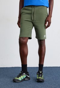 Olivenfarvede bomuldsblandede shorts med elastisk talje og snøre, prydet med et hvidt logo på det nederste ben. Båret med sorte sportssko.