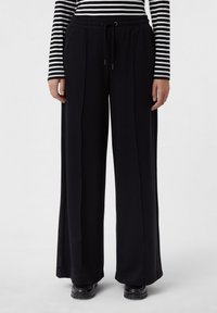 Pantalons larges noirs avec une taille à cordon de serrage, présentant des plis et une texture lisse. Associés à un haut à manches longues rayé.