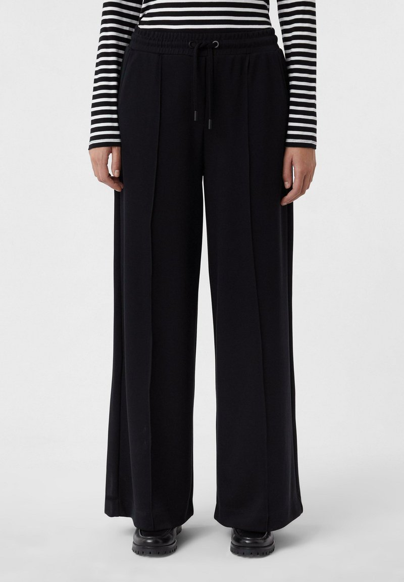 Pantalons larges noirs avec une taille à cordon de serrage, présentant des plis et une texture lisse. Associés à un haut à manches longues rayé.