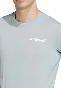 Camiseta atlética de corto azul claro con mangas cortas, cuello redondo, paneles laterales de malla y logo blanco "TERREX" en el lado izquierdo del pecho.