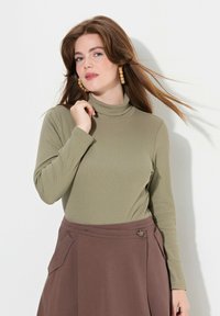 Maglione a collo alto verde oliva in materiale rigato, abbinato a una gonna marrone caratterizzata da una tasca laterale e dettagli con bottoni. Maniche lunghe, design aderente.