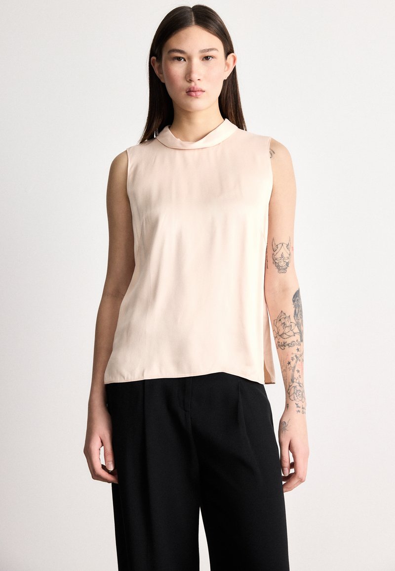 Blúzka bez rukávov v svetloružovej látke s hladkou textúrou, s okrúhlym výstrihom a bočnými rozparkami; v kombinácii s čiernymi nohavicami.