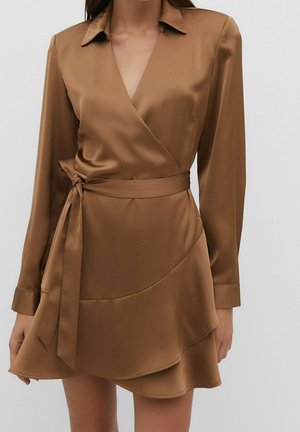 Kobieta w brązowej satynowej sukience kopertowej z długimi rękawami, kołnierzykiem i przewiązanym paskiem w talii na jednolitym jasnym tle.
