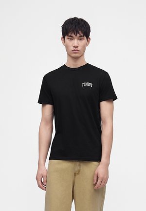 Jeune homme aux cheveux noirs courts portant un t-shirt noir de la marque Tommy et un pantalon beige sur un fond clair uni.