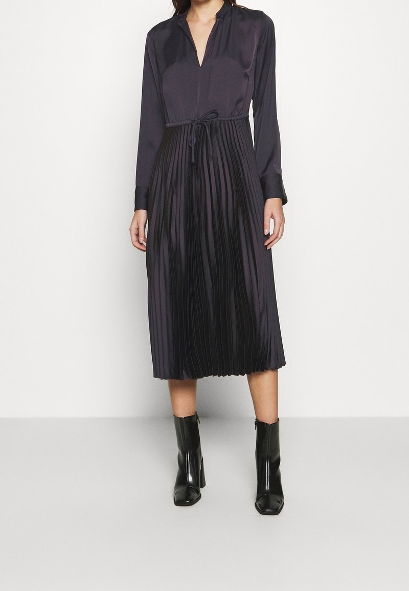 Femme portant une robe noire à manches longues avec une jupe plissée et une ceinture à la taille, associée à des bottines noires sur fond blanc.