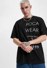 Melnas īsām piedurknēm T-krekls ar baltu "ROCA WEAR" rakstītu tekstu, dibināšanas gadu un pārāk lielu dizainu, kopā ar ziliem džinsu biksēm.