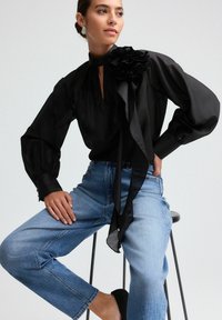 Sort langærmet bluse med høj hals og flæsedetaljer; parret med blå højtaljede denimjeans og sorte flade sko.
