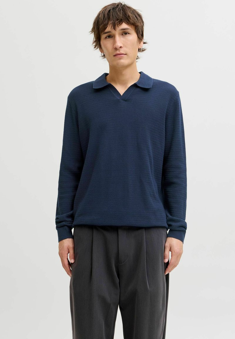 Polo bleu marine à manches longues avec un motif texturé, doté d'un col en V et d'un col rabattable, associé à un pantalon gris plissé.