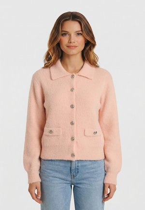 Femme portant un cardigan boutonné en tissu duveteux rose pâle avec un col et deux poches à l'avant, associé à un jean bleu clair.