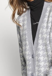 Cardigan grigio realizzato in materiale morbido, con motivo pied-de-poule, rifiniture a costine e due tasche anteriori. Chiusura con bottoni sul davanti.