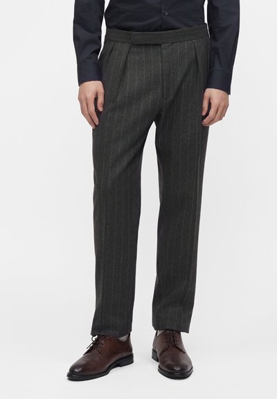 Homme portant un pantalon gris foncé à fines rayures et plis, une chemise bleu foncé et des chaussures habillées en cuir marron, debout devant un fond blanc.