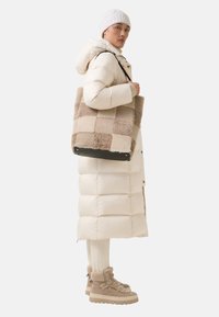 Persoon in een beige pufferjack, bijpassende gebreide muts, crèmekleurige broek en bontgevoerde wandelschoenen, met een beige patchwork tote bag.