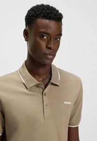 Beige pikétröja med en texturerad tyg, vit krage och treknapplås. Har en liten "HUGO"-logotyp på bröstet.