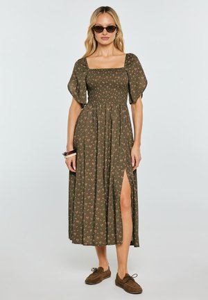 Kobieta ubrana w oliwkowo-zieloną midi sukienkę w kwiaty z bufkami i bocznym rozcięciem, brązowe loafersy, okulary przeciwsłoneczne i bransoletki, stojąca na jednolitym tle.