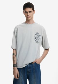 Világosszürke, rövid ujjú póló, laza szabással, grafikus logóval a bal melle részén. Puha pamut anyag.