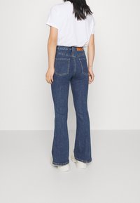 Nainen, jolla on yllään tummansiniset korkeavyötäröiset leveälahkeiset farkut, valkoinen T-paita ja valkoiset korkeat sneakerit, seisoo selin yksinkertaisella taustalla.