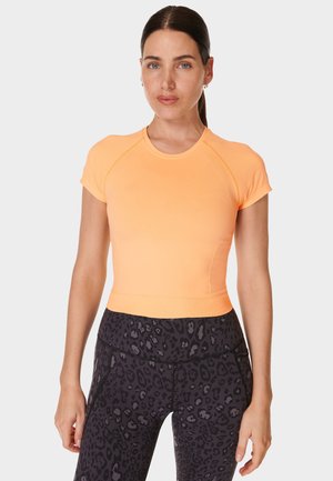 Woman with dark hair tied back wearing a short-sleeve orange athletic top and dark leopard print leggings, standing with arms relaxed.