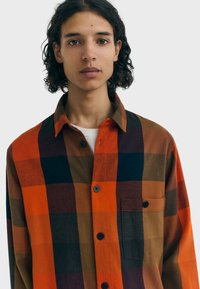 Chemise en flanelle présentant un grand motif tartan en orange, marine et marron. Fermeture à boutons, col et détail de poche poitrine visibles.