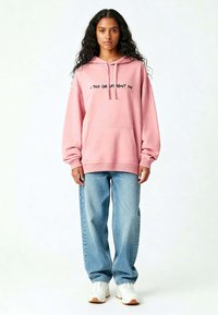 Roze capuchontrui met zwarte tekst, voorzak en een relaxte fit, gecombineerd met lichtblauwe wijde jeans en witte sneakers.