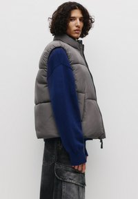 Grå puffervest med høj krave og lynlås, parret med en blå strikket trøje. Model iført mørke, strukturerede cargo-jeans.