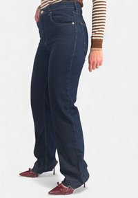 Jeans di denim blu scuro con una vestibilità a gamba dritta, caratterizzati da tasche frontali e cuciture dorate sottili. Indossati con scarpe con tacco alto color borgogna.