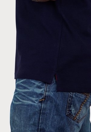 Marineblaues T-Shirt mit seitlichem Schlitz, kombiniert mit hellblauen Jeans. Jeans mit ausgefranstem Look und gelben Nähten an den Taschen.
