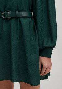 Dunkelgrünes Kleid aus strukturiertem Stoff, mit langen Ärmeln, einem taillierten Schnitt mit einem glatten Ledergürtel und einem lockeren Sitz.