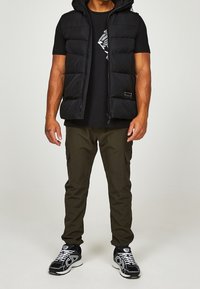 Černá prošívaná vesta s kapucí přes černé tričko s šedou grafikou, doplněná o olivové cargo kalhoty a černé tenisky.