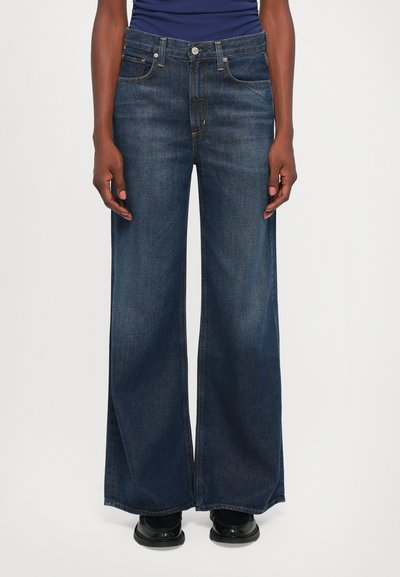 Persona che indossa jeans a gamba larga blu scuro, scarpe nere e una maglietta blu navy, in piedi contro uno sfondo bianco uniforme.