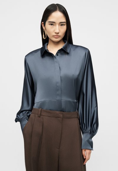 Γυναίκα με μακριά μαύρα μαλλιά φοράει μπλε σατέν μπλούζα με φουσκωτά μανίκια και καφέ ψηλόμεσο παντελόνι, στέκεται μπροστά σε απλό φόντο.