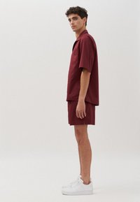Giovane in piedi di lato che indossa una camicia a maniche corte e pantaloni corti coordinati di colore bordeaux, abbinati a sneaker bianche su uno sfondo neutro.