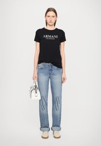 Svart T-skjorte med "ARMANI EXCHANGE"-logo i sølv, lyseblå jeans med logo mønster på bena, hvit håndveske, kremfargede sneakers.