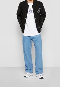 Man in een zwarte varsityjack, een wit graphic t-shirt met een duif, lichtblauwe jeans en witte sneakers, staand tegen een effen achtergrond.