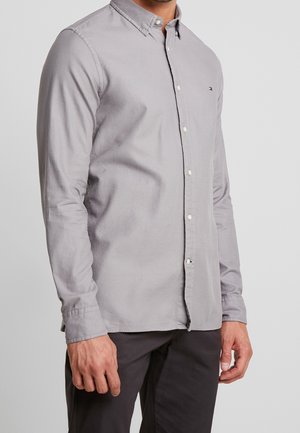 Hombre con camisa gris clara de manga larga con botones y pantalones negros, de pie frente a un fondo liso y claro.