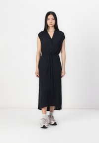 Abito midi blu navy con colletto, maniche a cappuccio, chiusura con bottoni e cintura in vita. La modella indossa sneakers rosa chiaro e calzini grigi.
