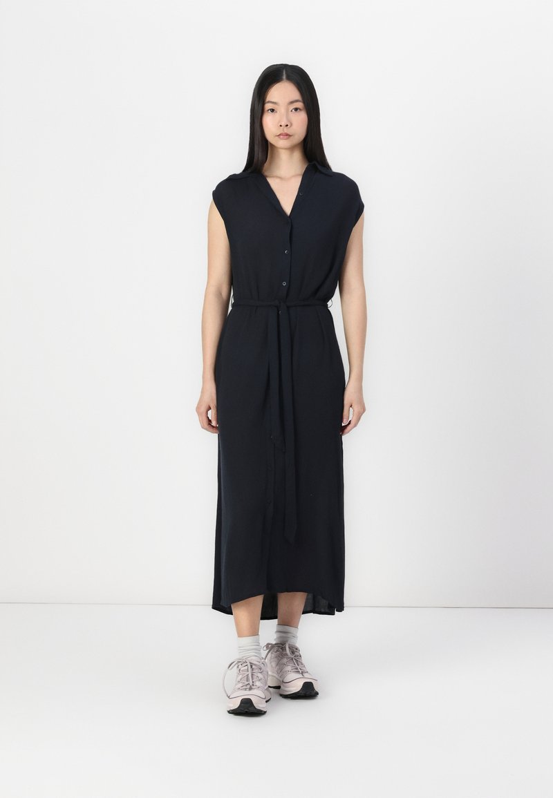 Abito midi blu navy con colletto, maniche a cappuccio, chiusura con bottoni e cintura in vita. La modella indossa sneakers rosa chiaro e calzini grigi.