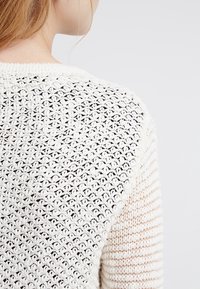 Pull en crochet avec un motif ajouré sur le haut du dos et une texture côtelée unie sur la partie inférieure, principalement en couleur crème.