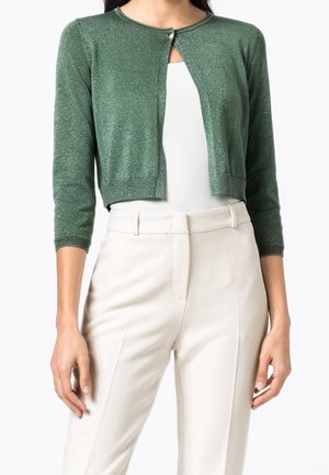 Mujer llevando un cárdigan corto verde brillante con mangas tres cuartos sobre una blusa blanca y pantalones beige de cintura alta.