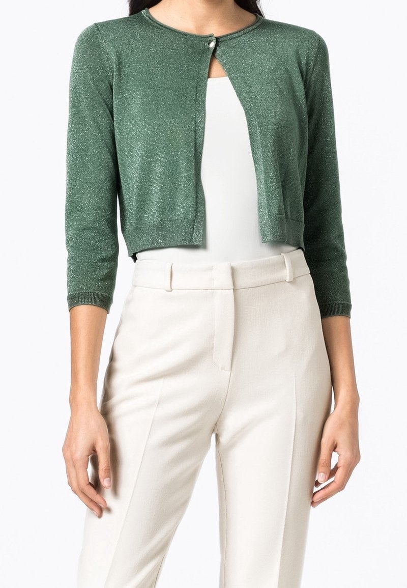 Femme portant un cardigan court vert scintillant à manches trois-quarts sur un top blanc et un pantalon beige taille haute.