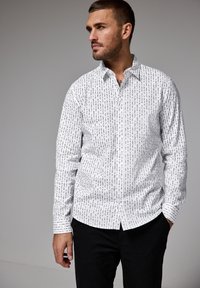 Bílá košile na knoflíky s černým potiskem, s klasickým límcem, dlouhými rukávy a manžetami na knoflíky, v kombinaci s černými kalhotami.